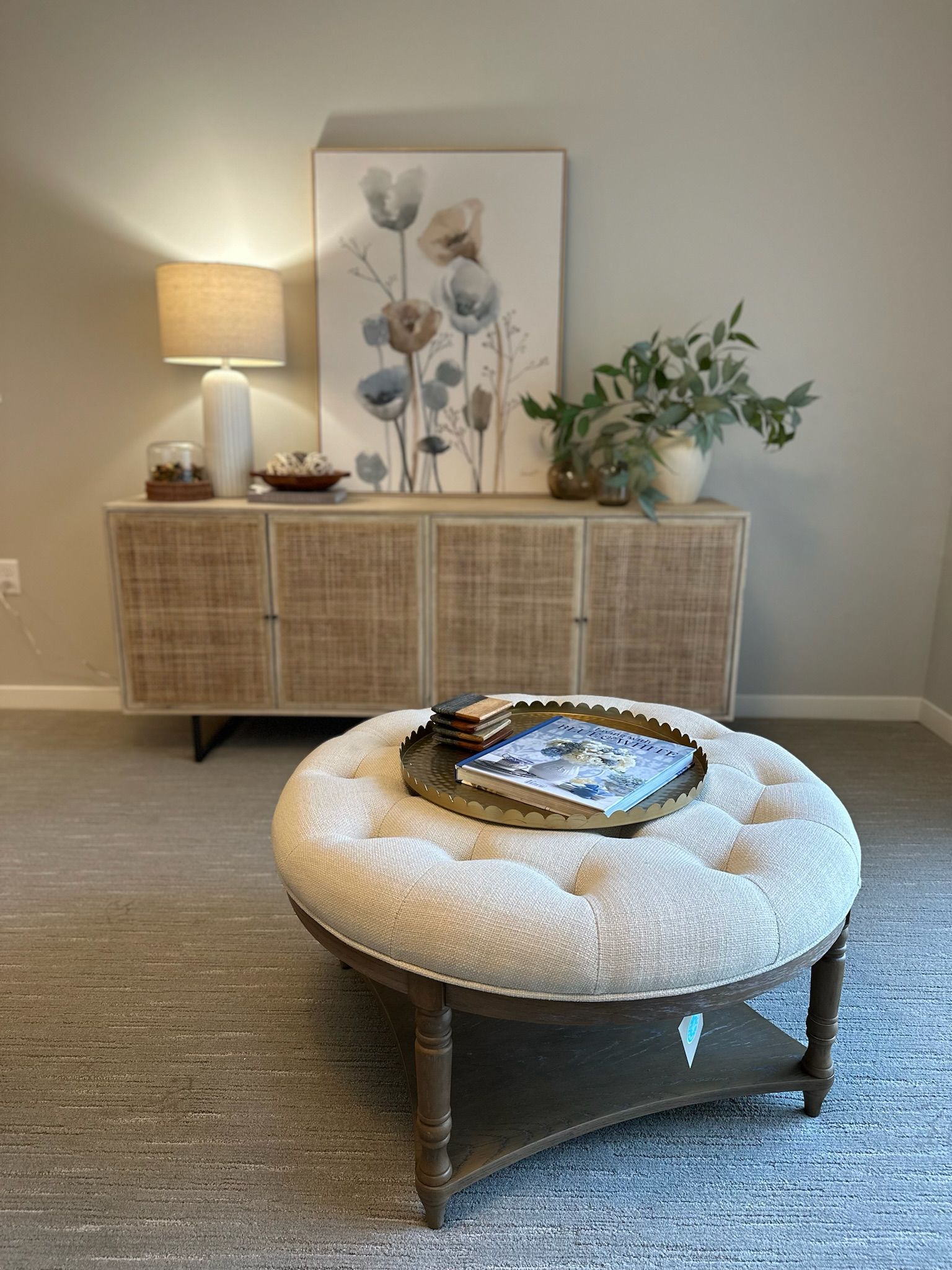 A tufted, cream-colored ottoman with a tray sits before a woven sideboard topped with a lamp, art, and a potted plant.