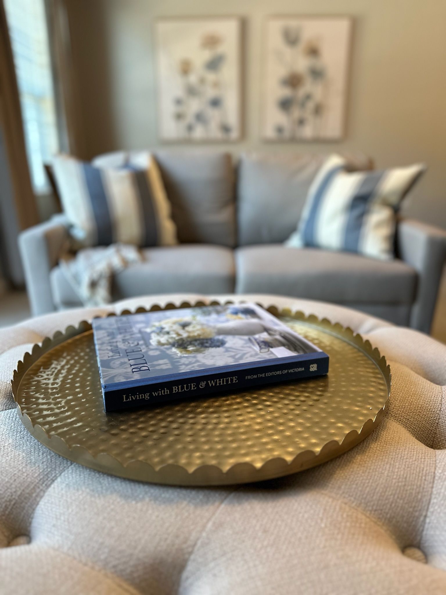 A textured gold tray holds a book on a tufted ottoman in front of a grey sofa with striped pillows and framed wall art.
