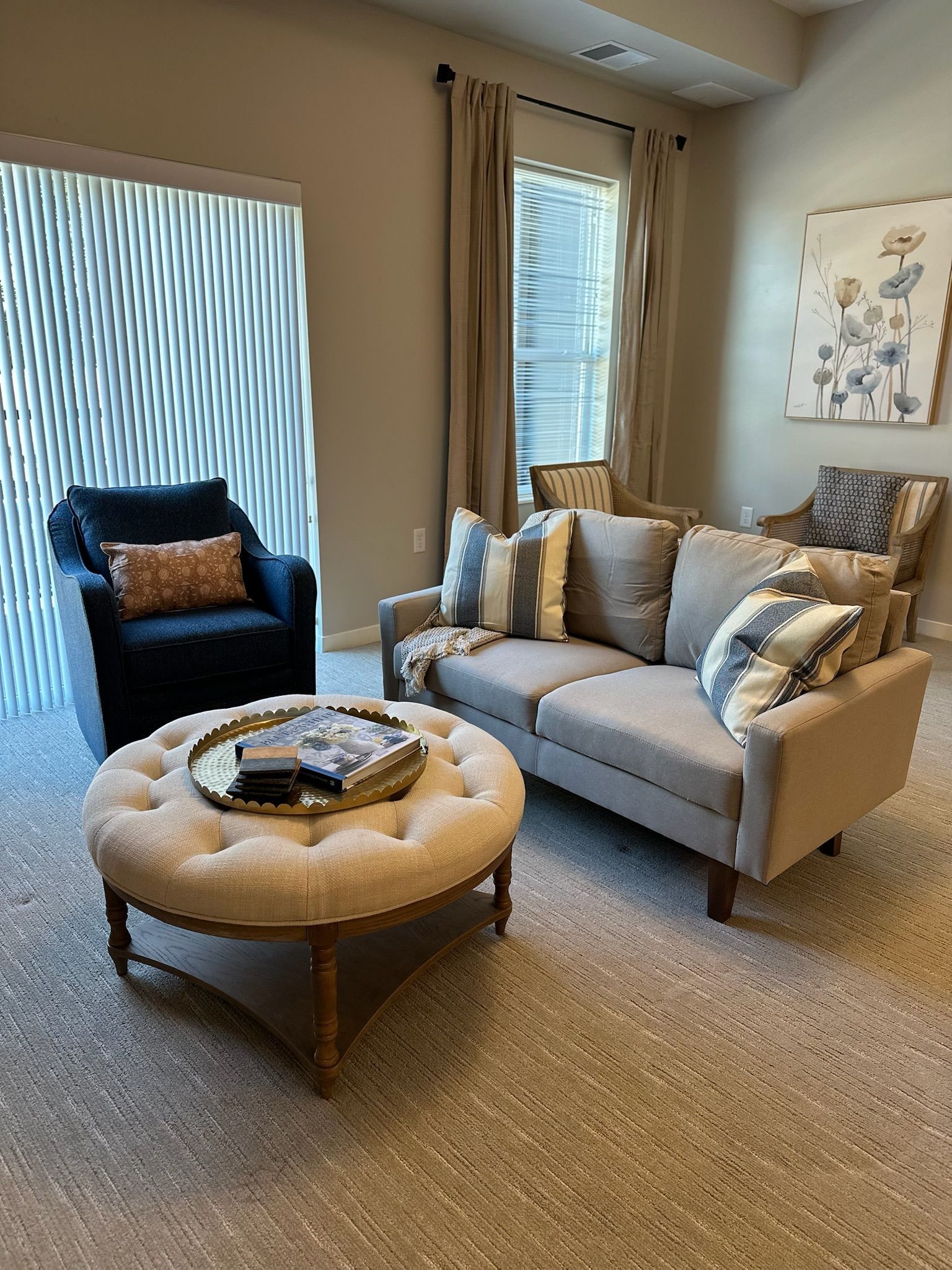 A modern living area featuring a beige sofa, dark blue armchair, circular tufted ottoman, and light-filled windows.