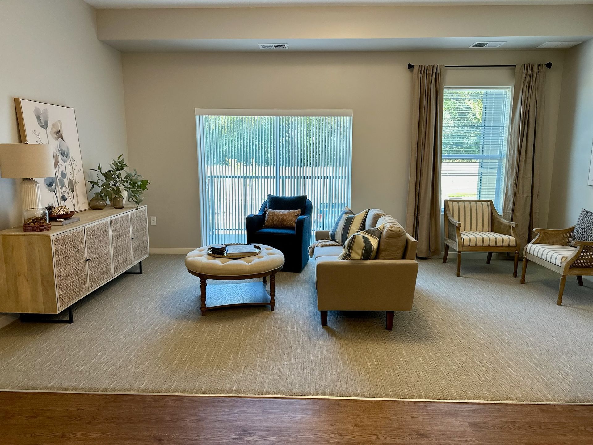 A living room with a beige sofa, dark armchair, round ottoman, light wood console, and two side chairs on neutral carpet.