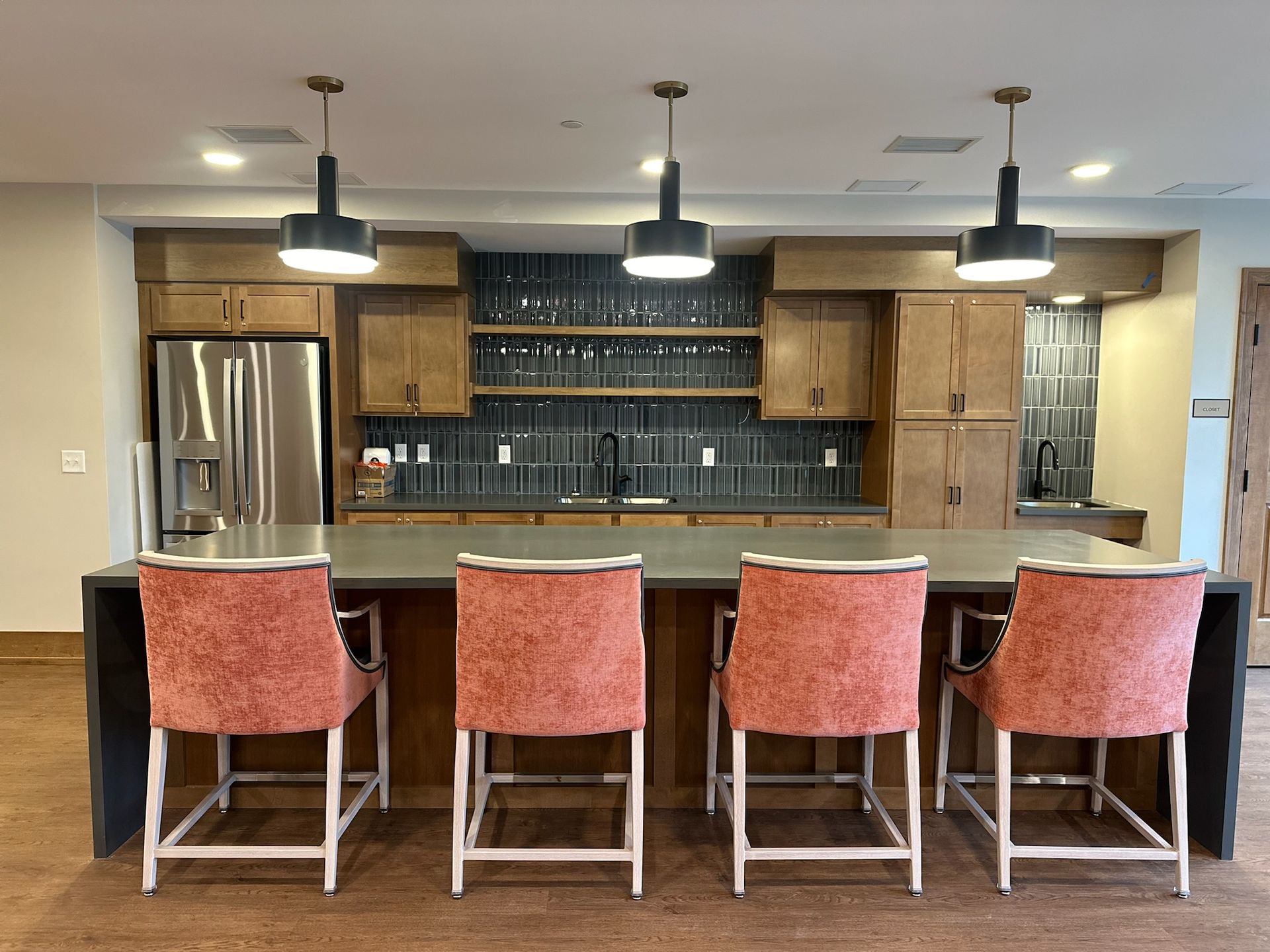 A modern kitchen with a large island featuring four pink chairs, wooden cabinets, and three hanging pendant lights.