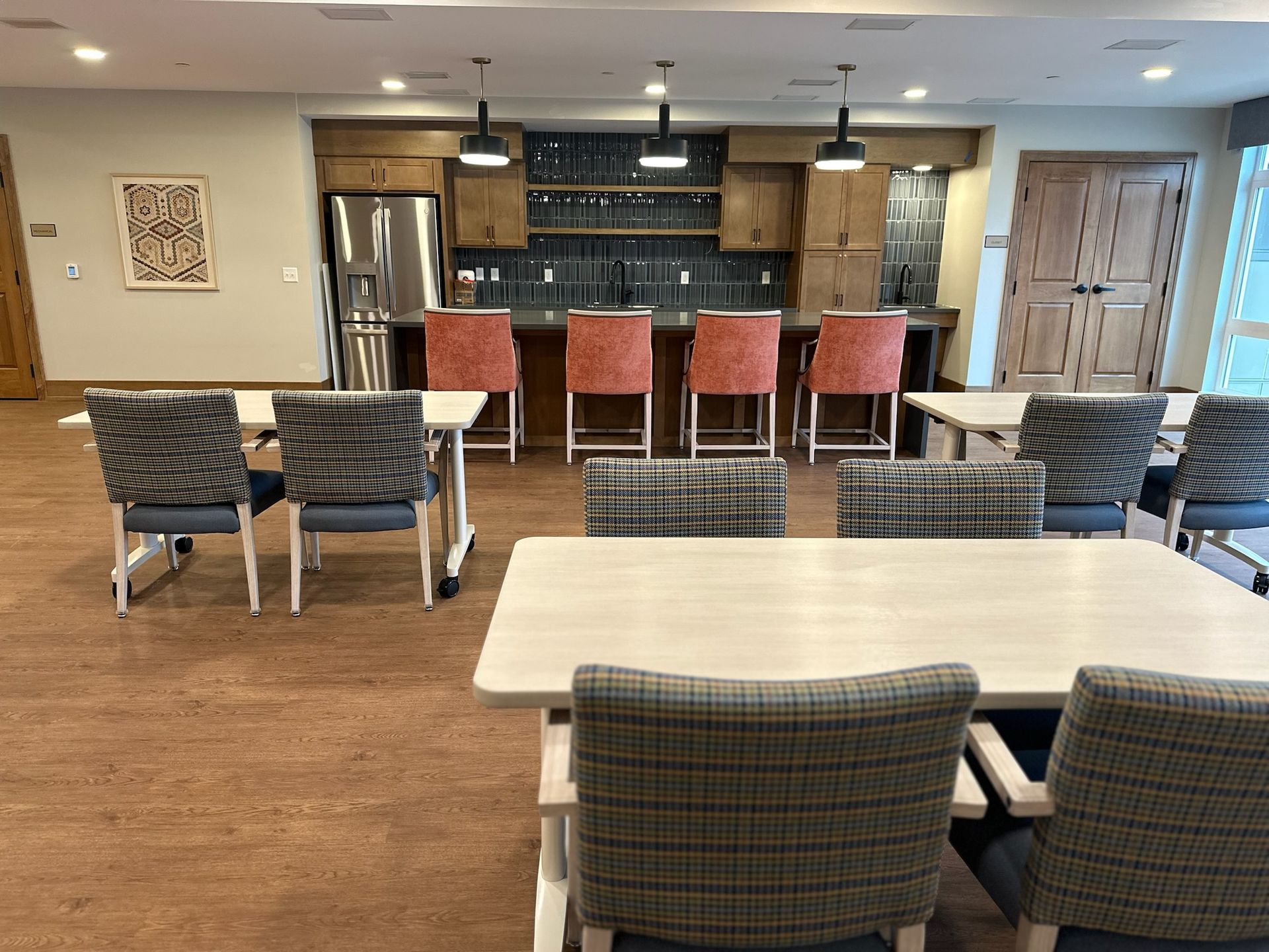 A community room with tables, chairs, and a bar area with four coral-colored stools and pendant lighting.