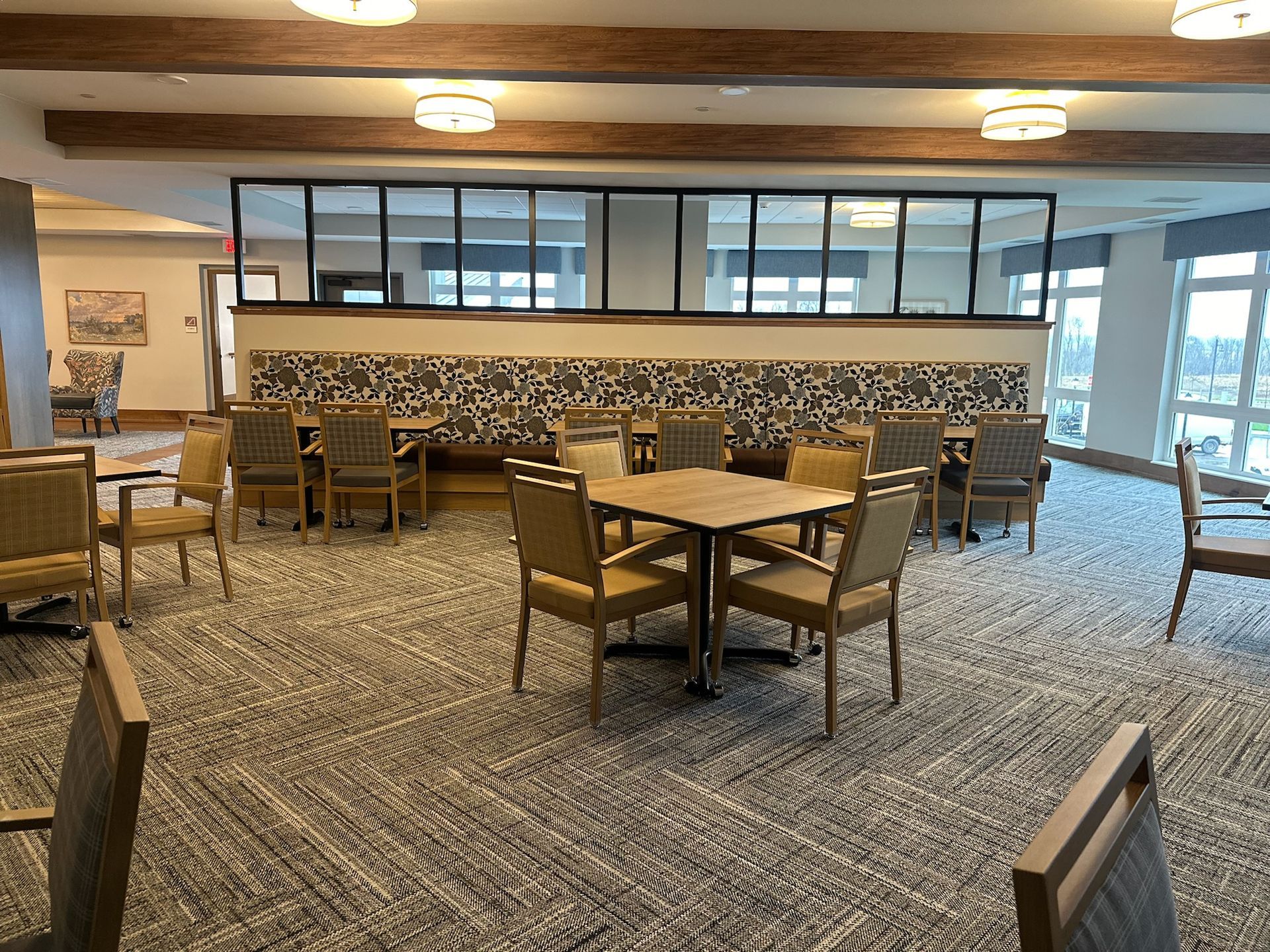 A bright, spacious dining area with several square tables and chairs, wood ceiling beams, and large windows.