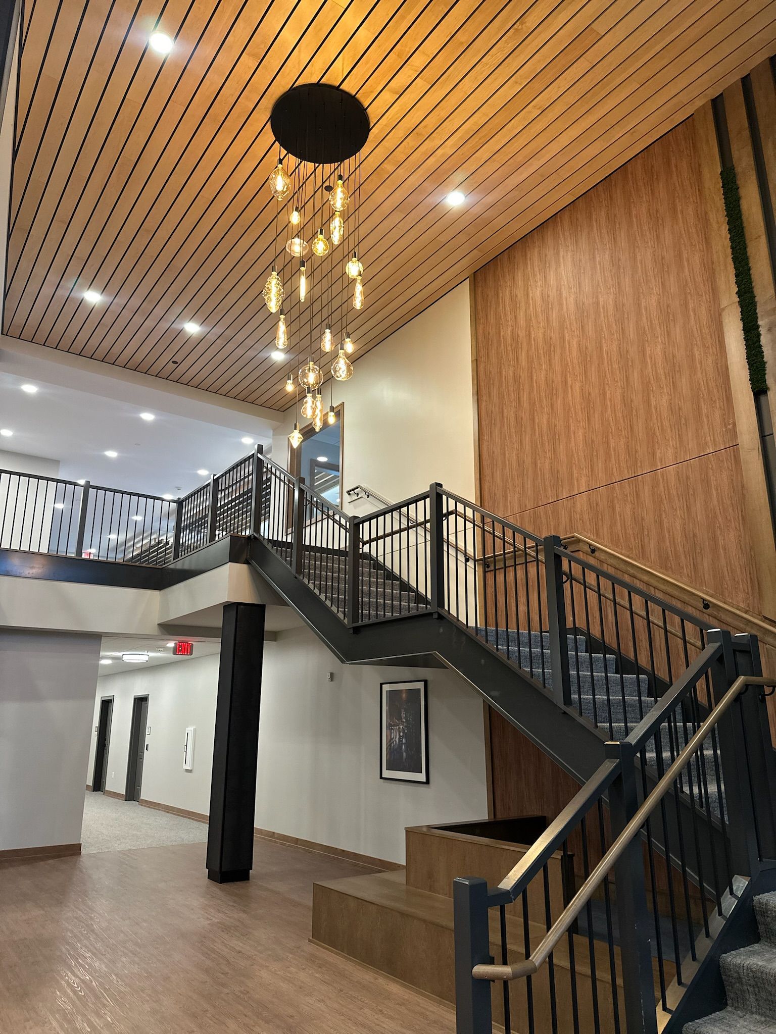 A modern building interior featuring a tiered wooden staircase, a metal railing, a tall wood-paneled wall, and a chandelier.