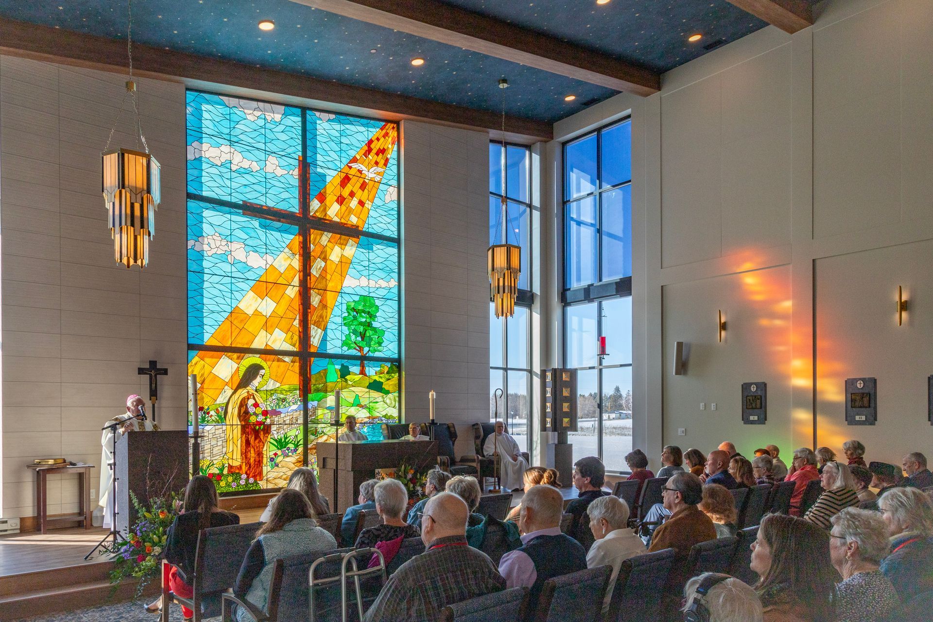 People sit in a church during a service, facing a large, colorful stained-glass window of a figure.