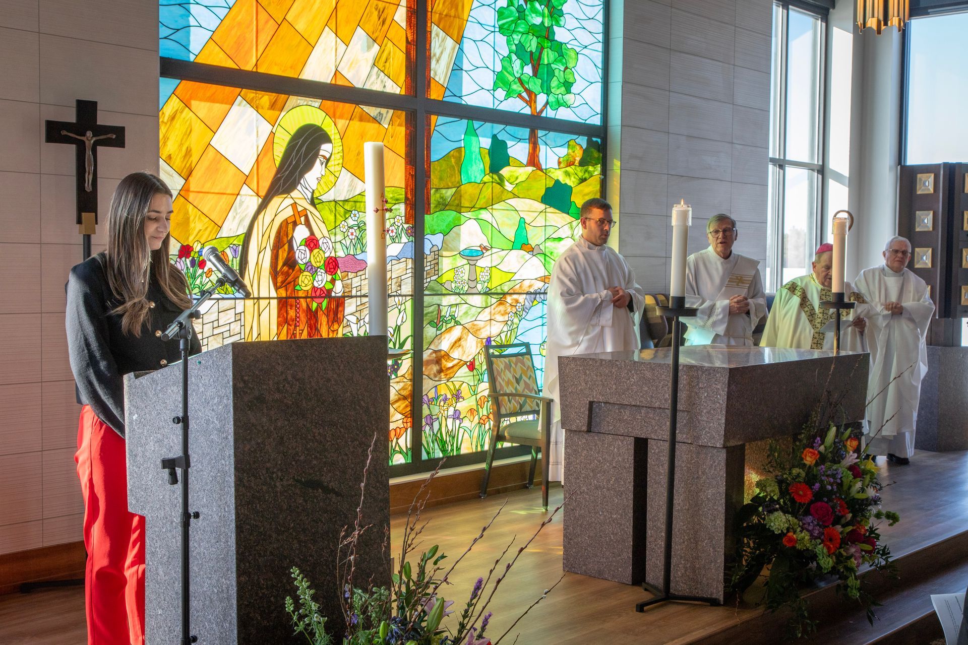 A person stands at a lectern reading during a church service, with clergy and a colorful stained glass window behind them.