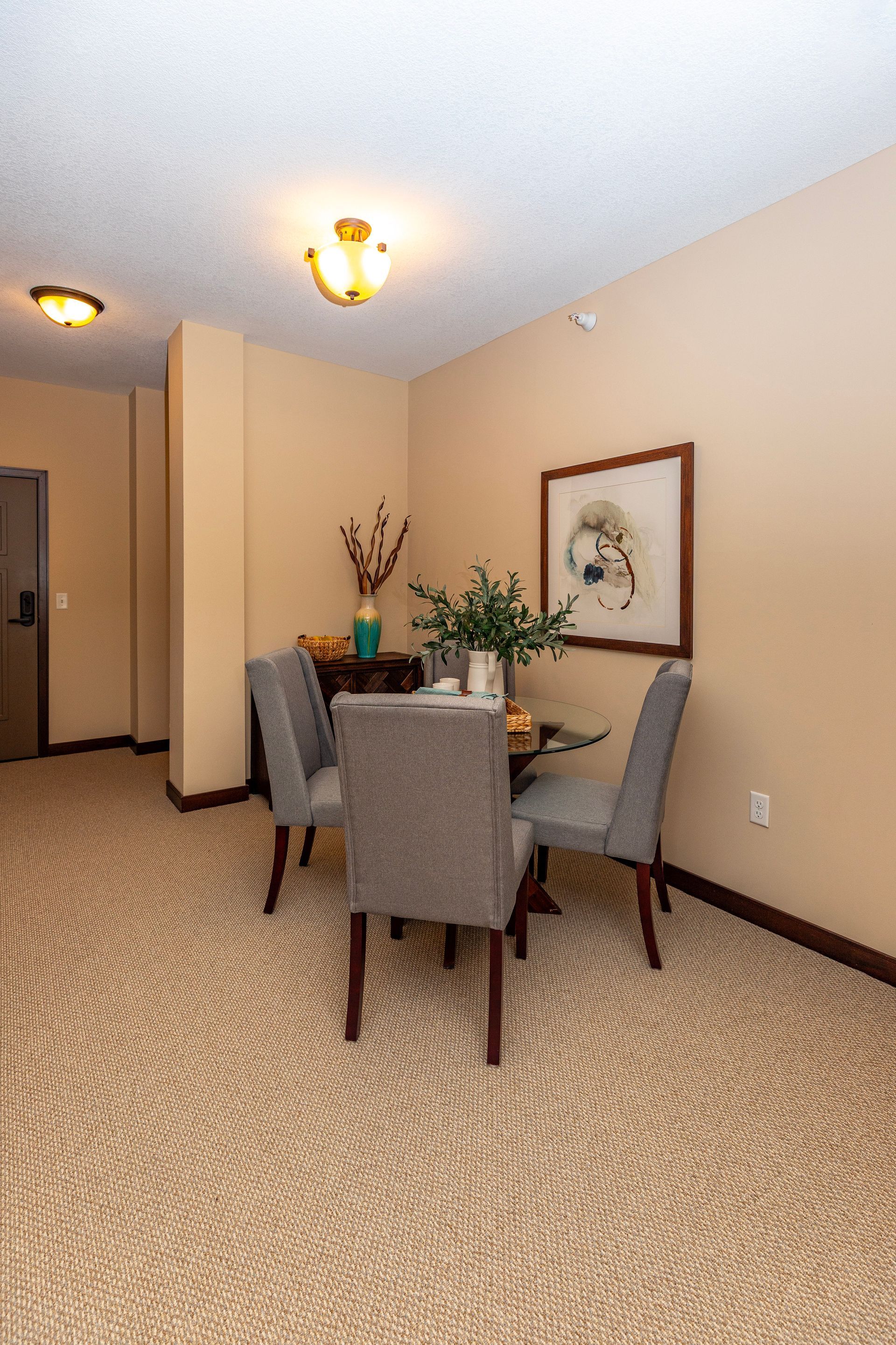A dining set with four grey upholstered chairs and a glass table sits on a tan patterned carpet in a neutral-toned room.