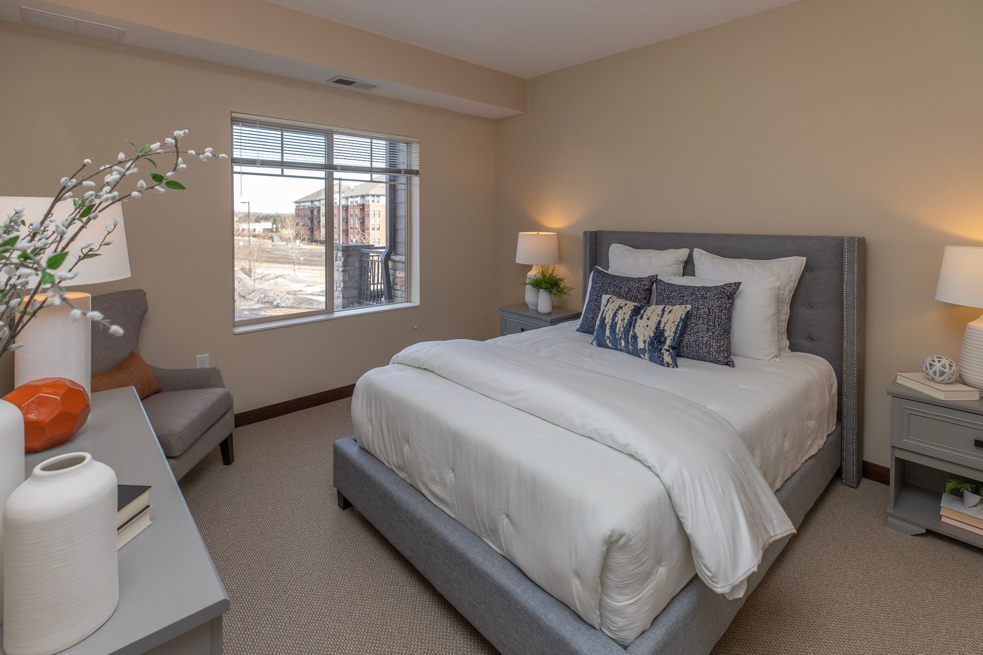 A light-colored bedroom features a grey upholstered bed, a grey chair by a window, and two bedside tables with lamps.