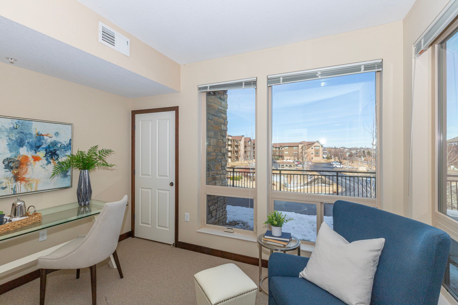 A brightly lit home office with a glass desk, white chair, blue armchair, and a large window overlooking a snowy area.