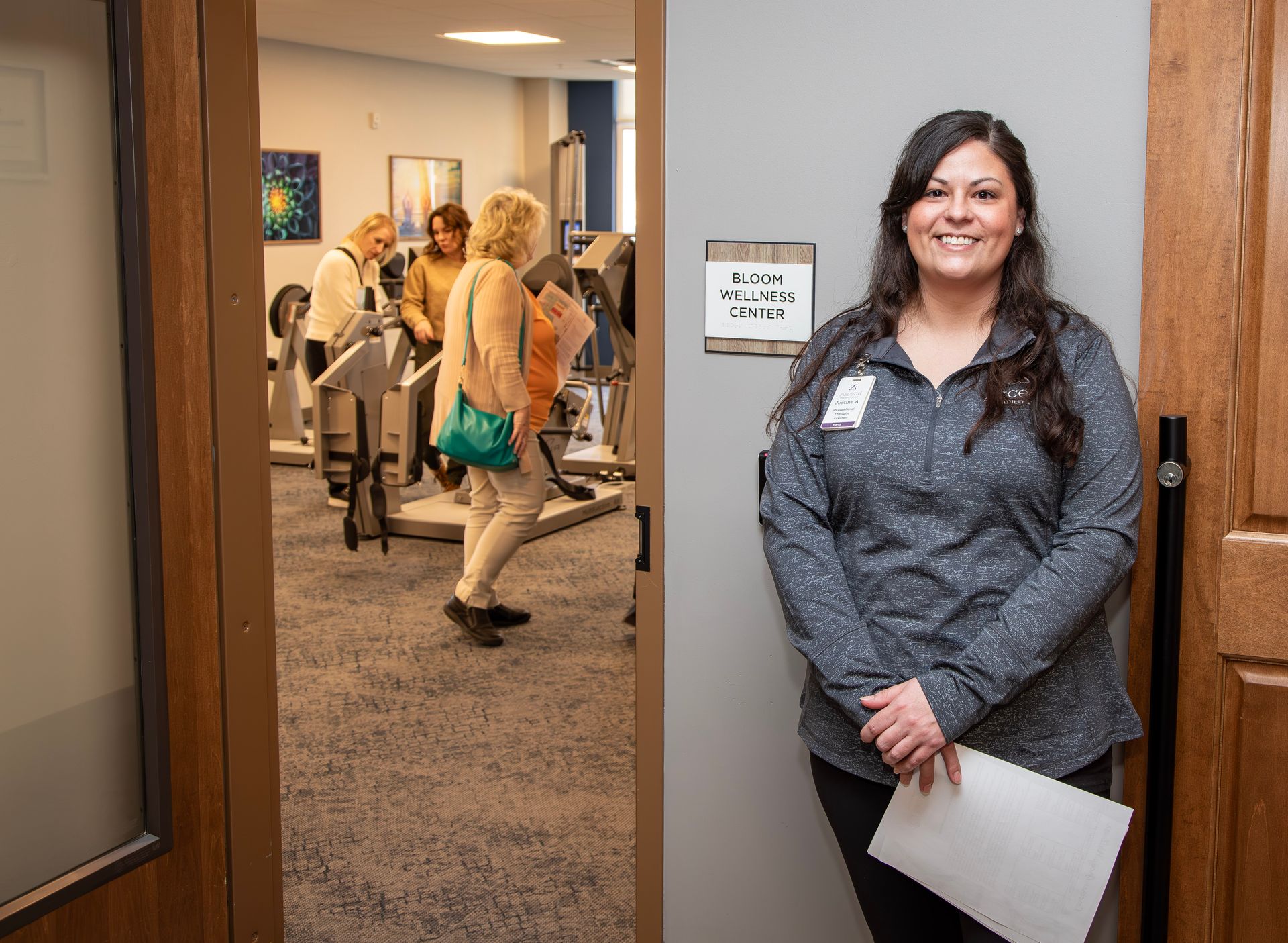 A smiling staff member stands in a doorway; inside, three people exercise on equipment in a carpeted physical therapy room.