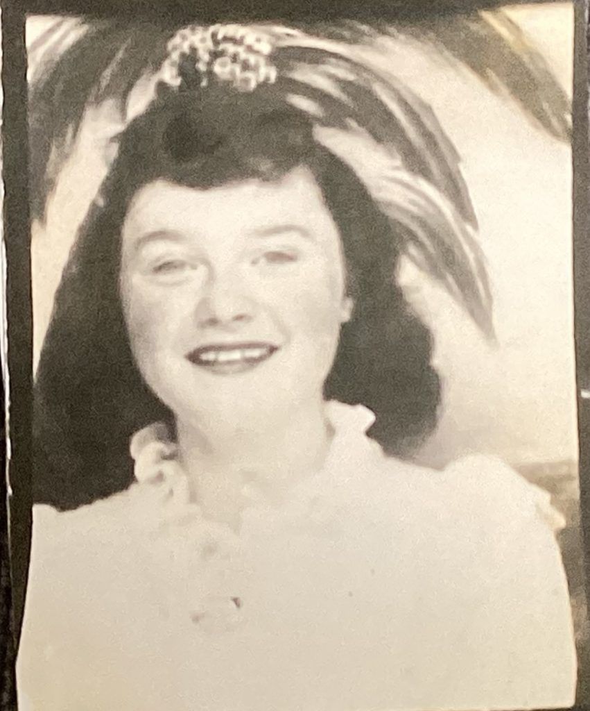 A black-and-white portrait of a person smiling, wearing a blouse with a ruffled collar and a large feathered hairpiece.