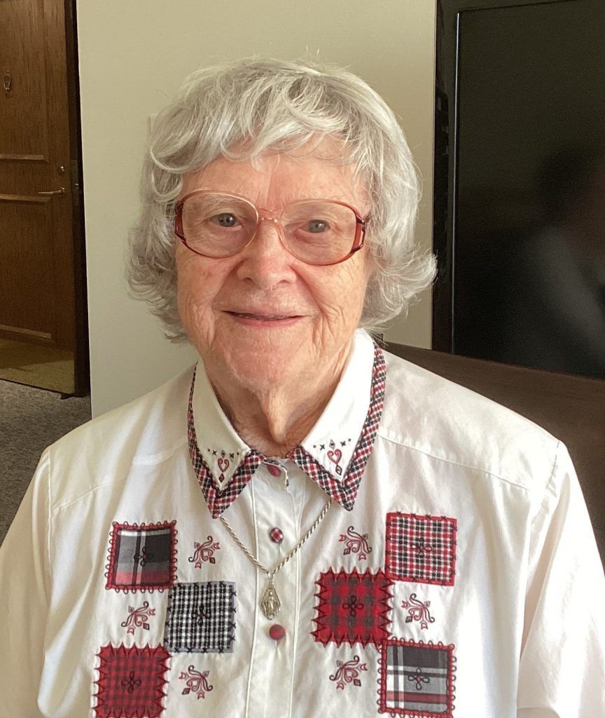 A person wearing glasses and a white button-down shirt with red and black plaid patch accents smiles at the camera.