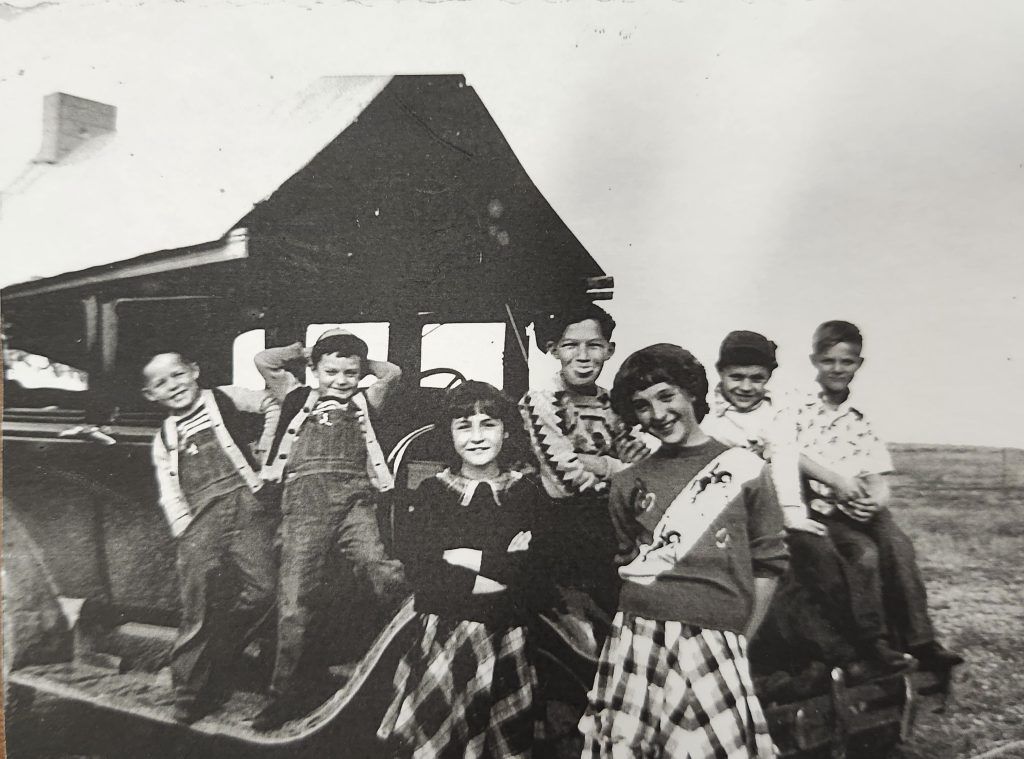 Seven children stand and sit on the hood and bumper of a vintage car in an open field, smiling at the camera.