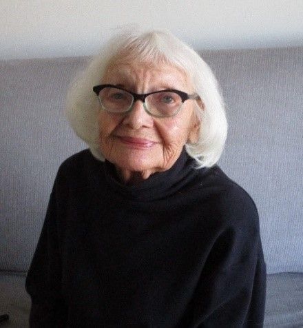 A person with white hair and dark-rimmed glasses wearing a black turtleneck, sitting in front of a light gray wall.