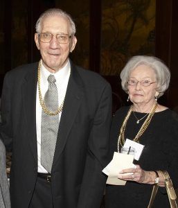 A person in a dark suit and tie with a gold chain stands next to a person in a black top wearing multiple gold necklaces.