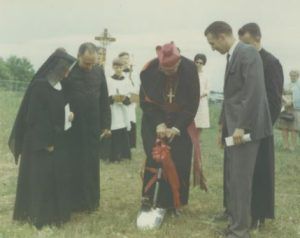 A bishop in vestments performs a ceremonial groundbreaking with a shovel, surrounded by religious and lay observers.