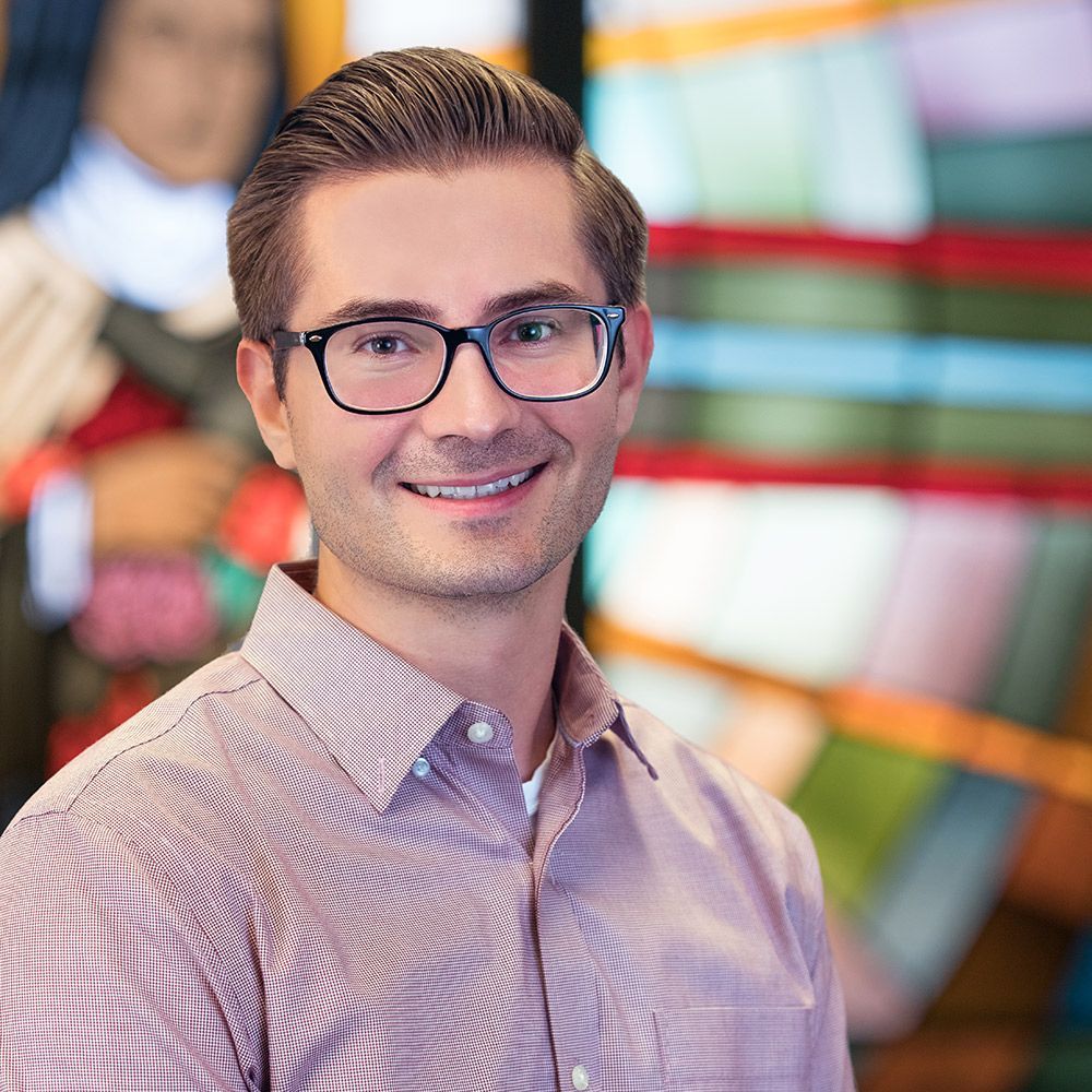 A professional headshot of a person with short brown hair and glasses, wearing a patterned shirt, smiling at the camera.