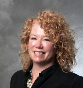 A person with curly blonde hair and a pearl necklace smiling against a grey background.