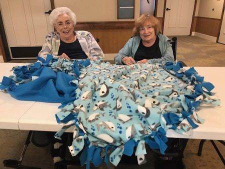 Two people smile while crafting a blue, shark-patterned fleece tie blanket on a table in a community room.
