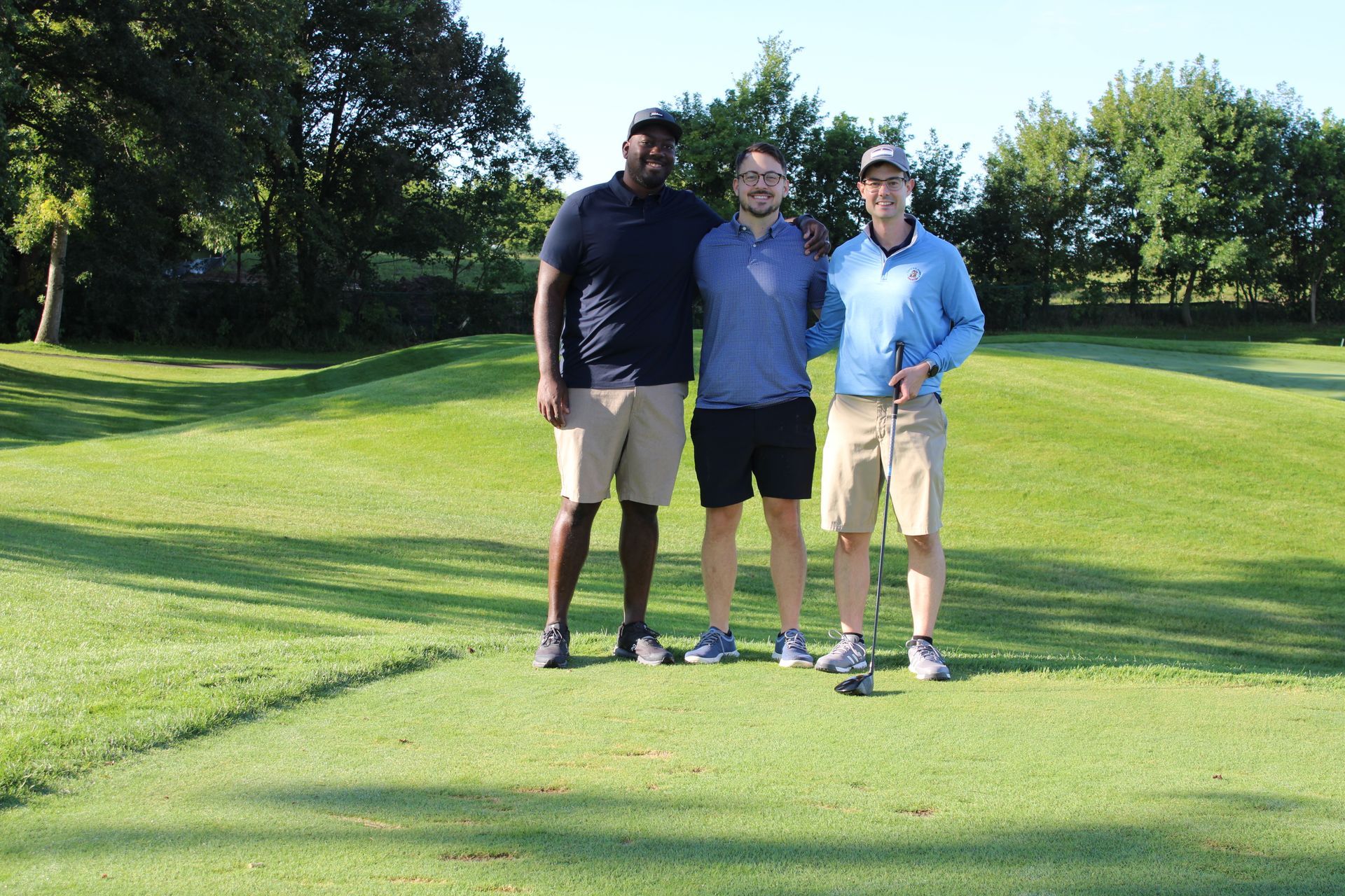 Three people standing together on a sunny golf course, smiling and posing for a group photo.