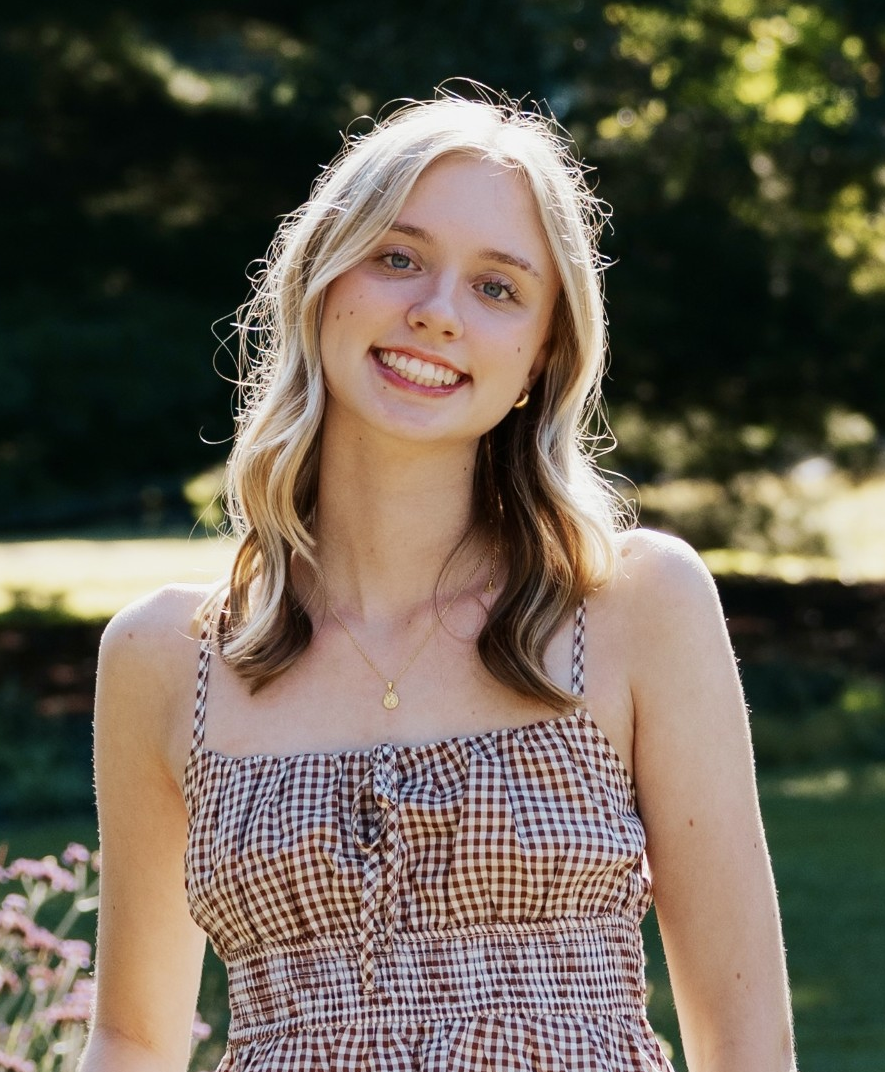 Smiling person in a brown checkered dress standing outdoors in sunlight with blurred greenery behind.