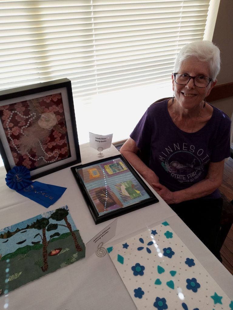 A person smiles beside a table displaying framed artwork, including a piece with a blue ribbon, in front of a window.