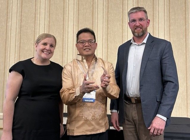 Three people stand together, with the center individual holding an award, against a light beige vertical-paneled wall.
