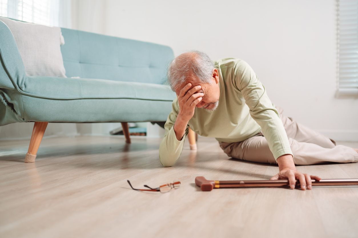 An older adult sits on the floor after a fall, holding their head with one hand near their glasses and walking cane.