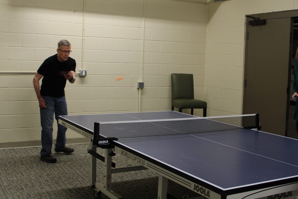 A person in a black shirt and jeans plays table tennis in a room with light-colored brick walls.