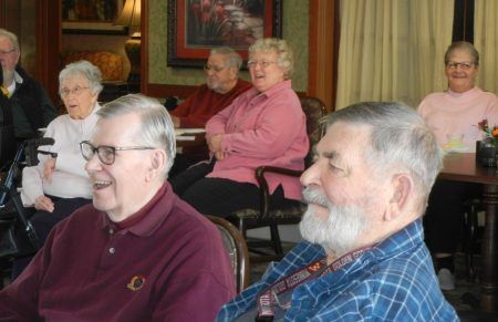 A group of people sitting at tables in a room, smiling and listening during a gathering.