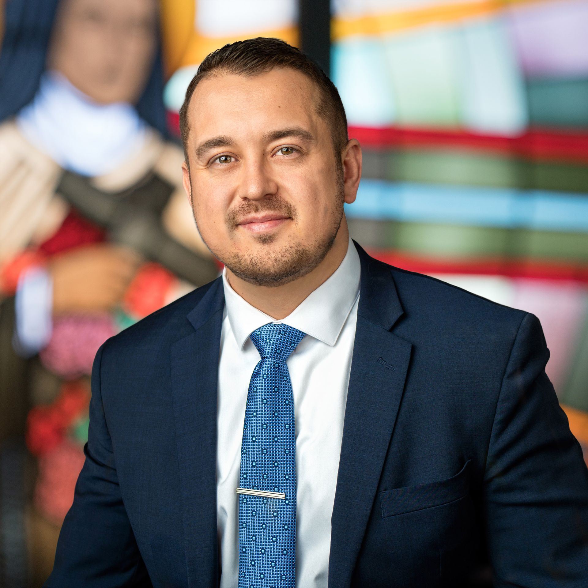 Headshot of a professional in a navy suit, white shirt, and blue patterned tie, standing before a blurred colorful backdrop.