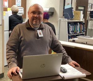 A person smiles at a laptop on a standing desk in an office setting with shelves and documents in the background.