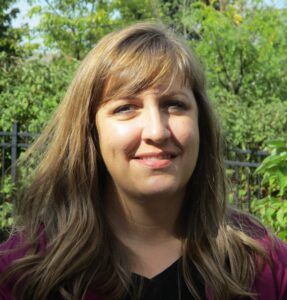 A woman with light brown hair smiles gently in front of a blurred green, leafy garden setting.