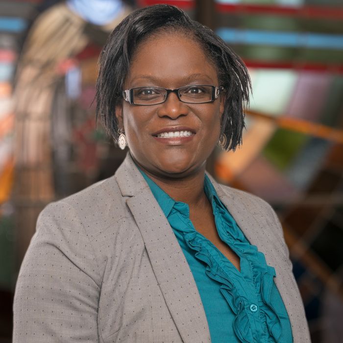 A person wearing glasses, a teal ruffled blouse, and a grey blazer, smiling warmly against a blurred, colorful background.