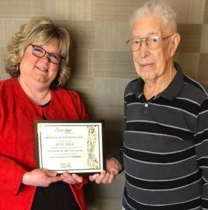A woman in a red jacket presents an award plaque to an older man in a striped shirt against a beige tiled wall.