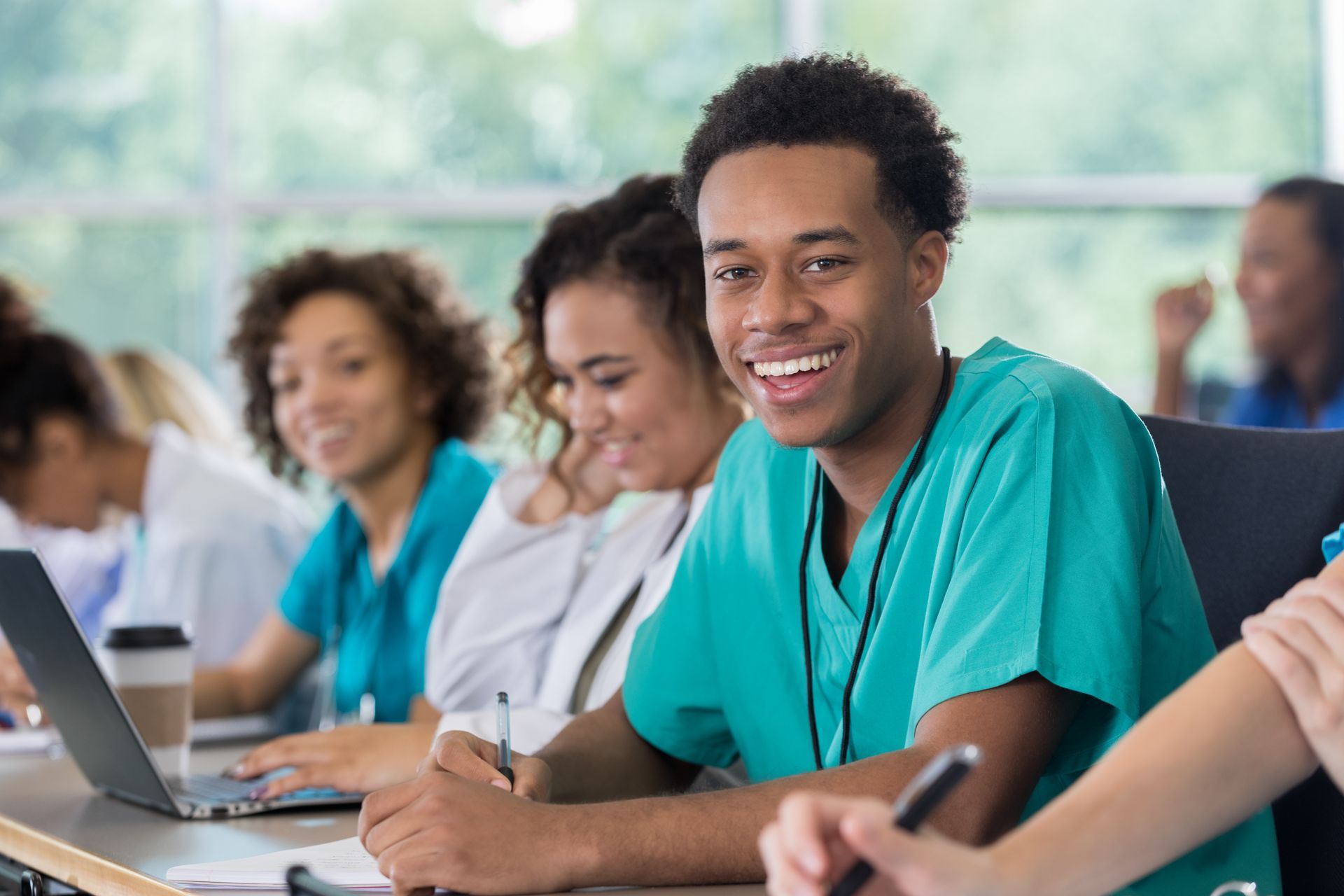 A smiling student in teal scrubs writes at a desk in a classroom, with other students studying in the background.