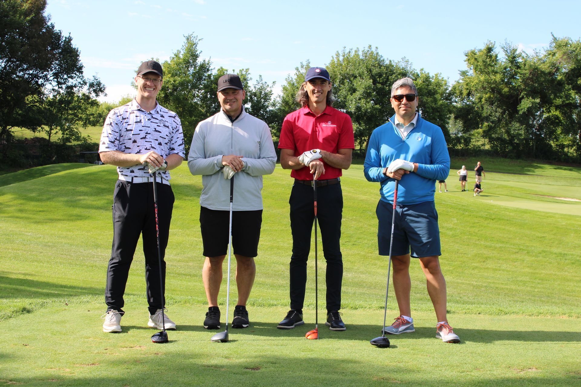 Four people stand in a line on a sunny golf course, each holding a golf club and smiling toward the camera.
