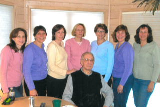 Seven people stand in a row behind a seated man in an indoor setting with wood-paneled walls.
