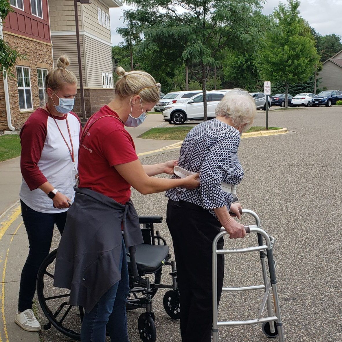 Two healthcare workers in masks assist an individual using a walker and wheelchair in a parking lot.