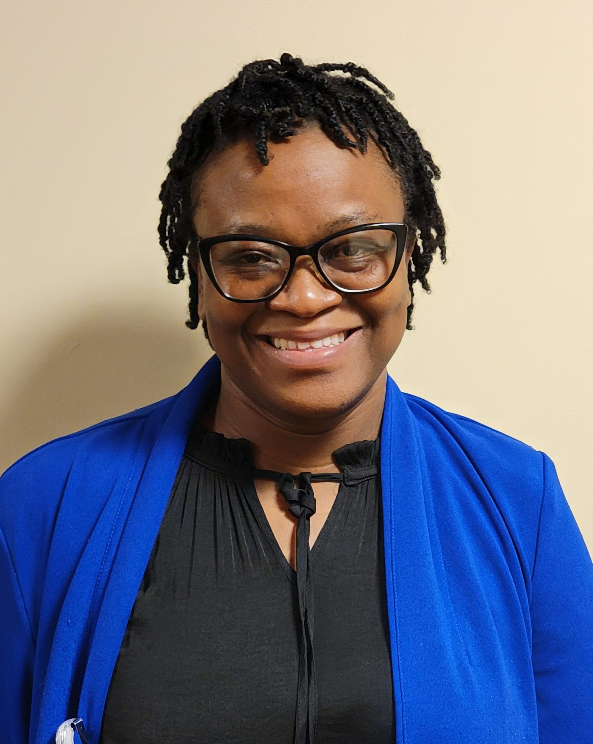 A person with short, textured hair wearing dark glasses and a royal blue cardigan over a black top, smiling at the camera.