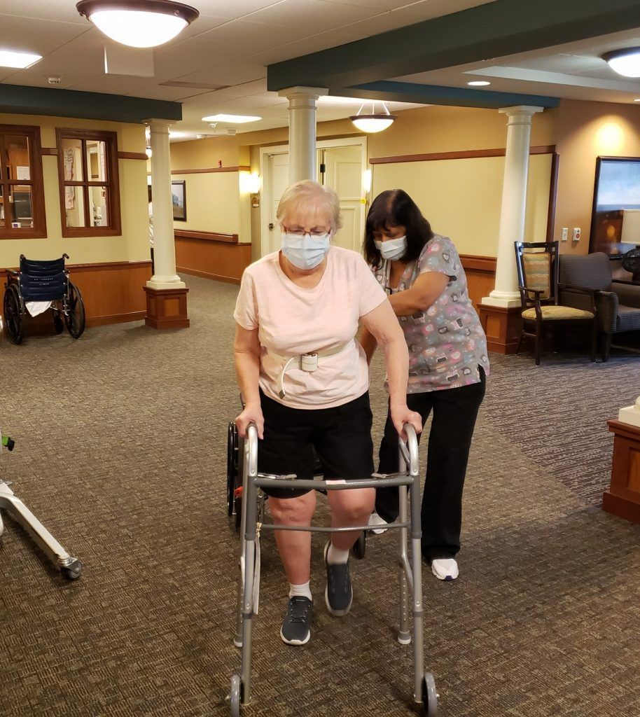 A caregiver assists a person with a walker in an assisted living facility hallway.