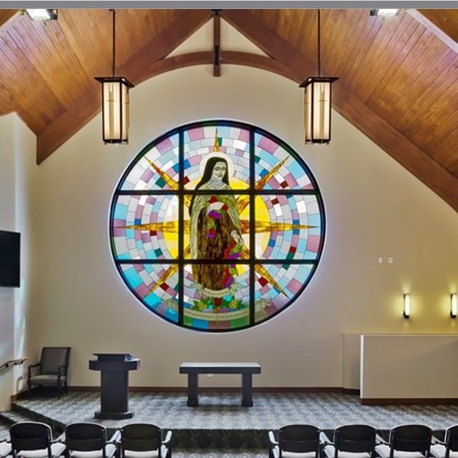 A modern chapel interior featuring a large, colorful circular stained-glass window of a religious figure above an altar.
