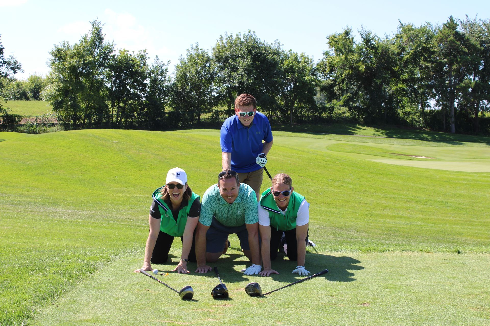 Four people pose on a golf course; three kneel in a row while one stands behind, all smiling with golf clubs on the grass.