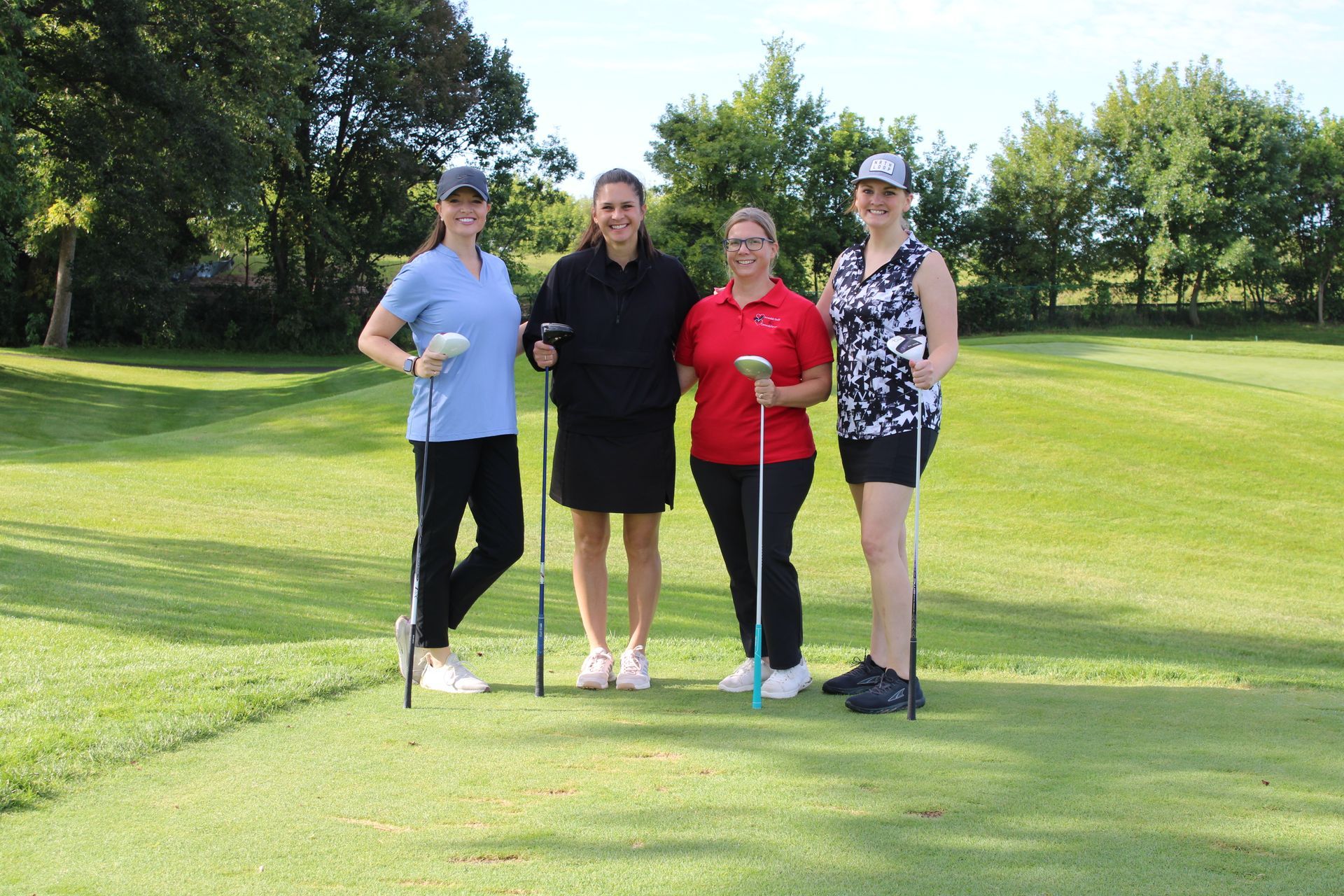 Four people stand on a grassy golf course, each holding a golf club, with trees and a bright sky in the background.