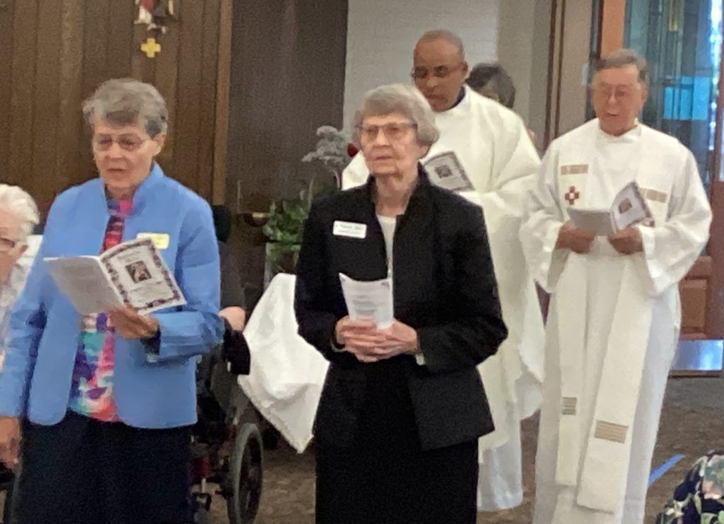 Four individuals, two in clergy attire, standing in a church-like setting holding programs and looking forward.