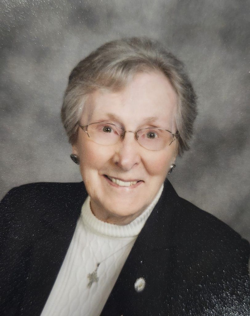 A headshot of a smiling person with gray hair wearing glasses, a white sweater, and a black jacket against a gray backdrop.
