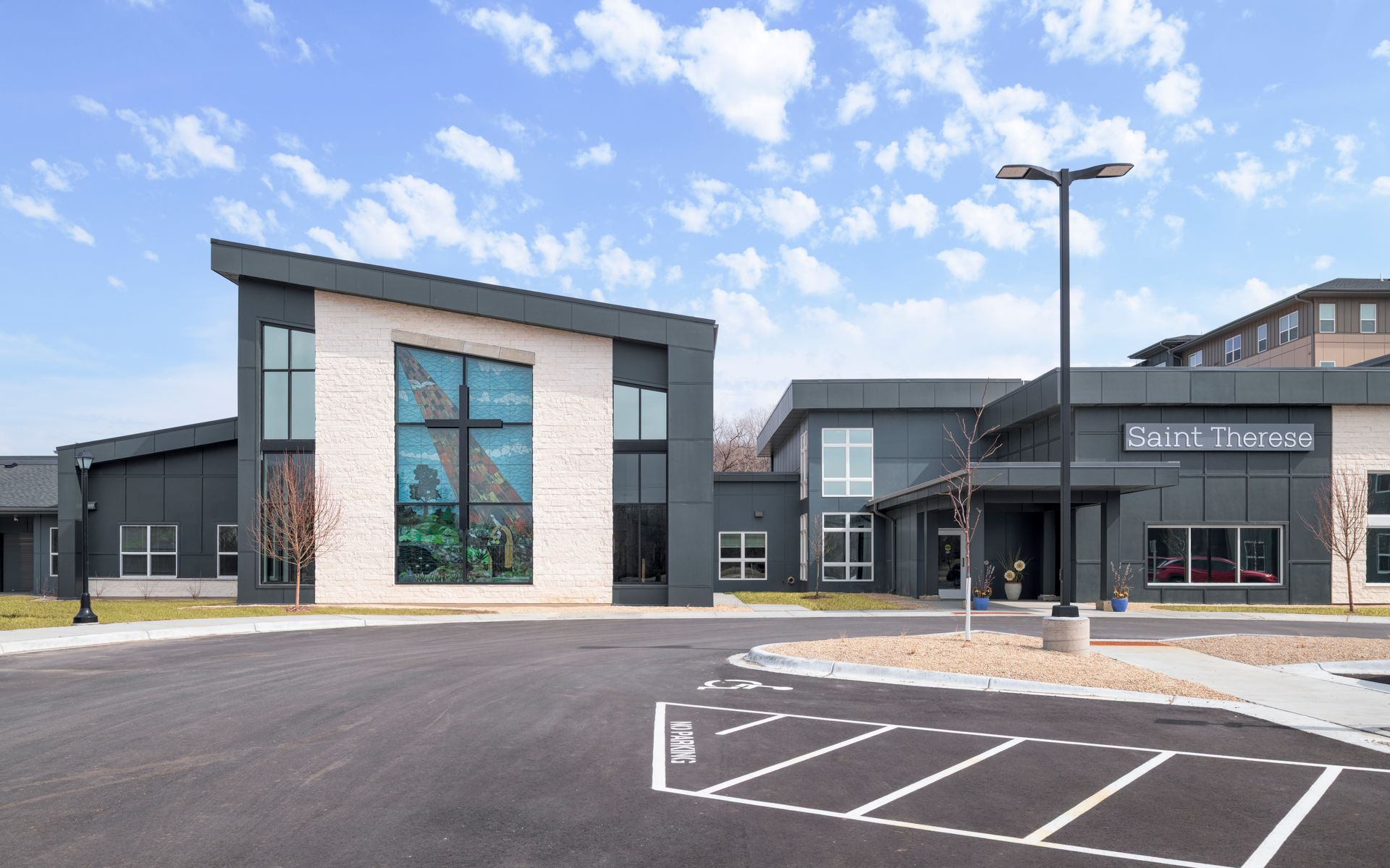 Modern church building with a light stone facade, dark metal panels, and a large front window featuring a cross.