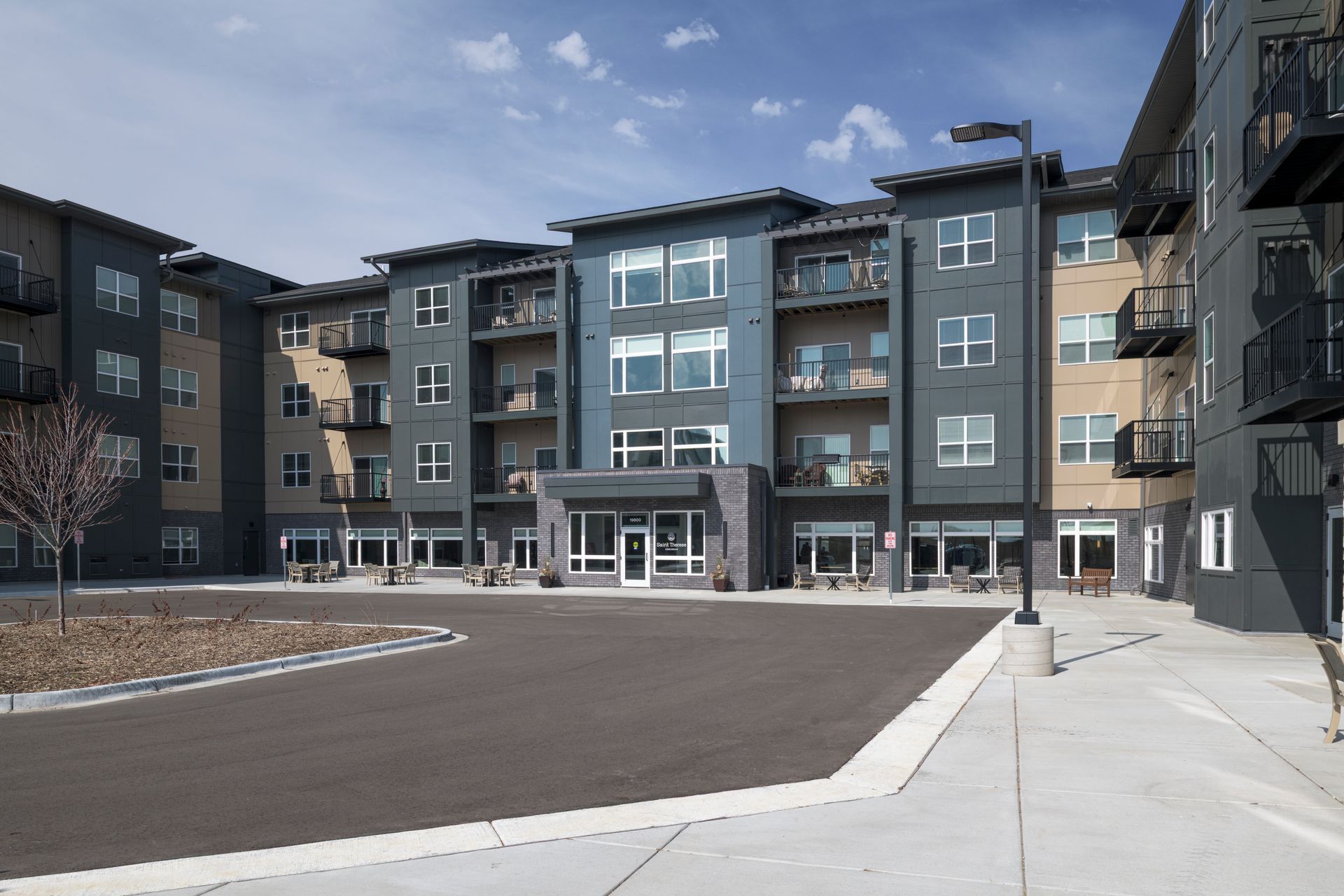 A modern, multi-story apartment building with dark blue and beige exterior panels, balconies, and a paved courtyard.