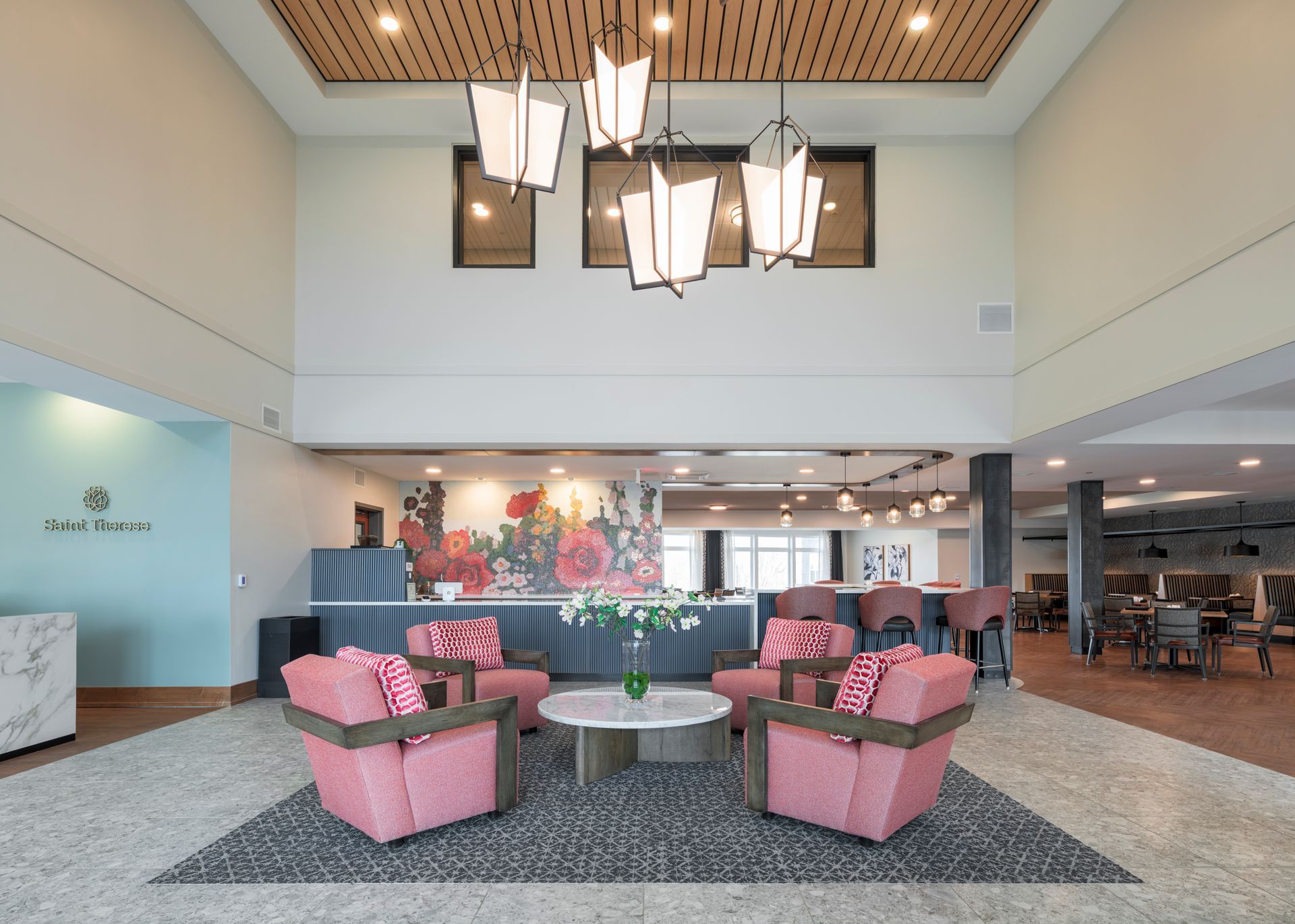 Spacious hotel lobby with pink armchairs, a central coffee table, a floral wall mural, and modern hanging light fixtures.