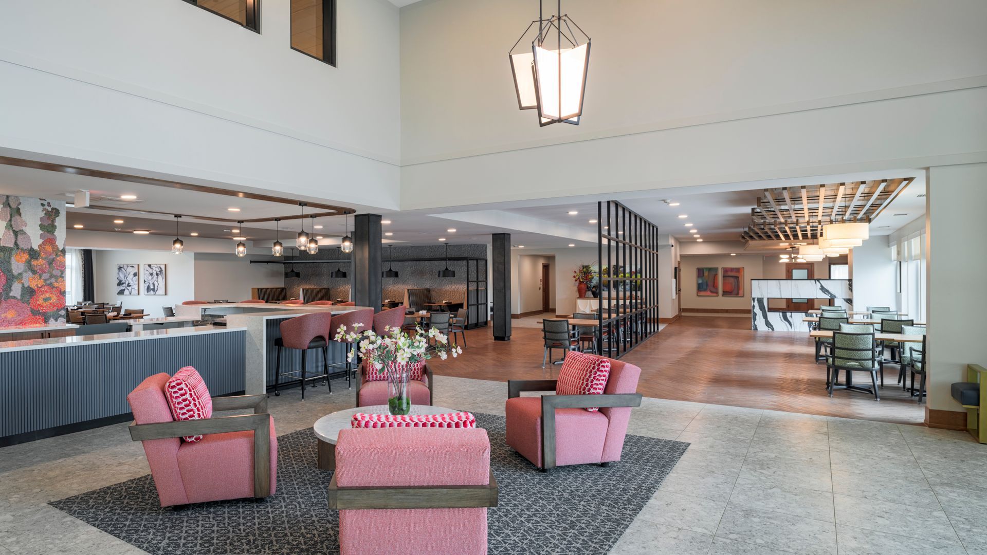 A modern, open-plan lobby with three pink armchairs around a circular table on a dark rug, with a bar and seating area.
